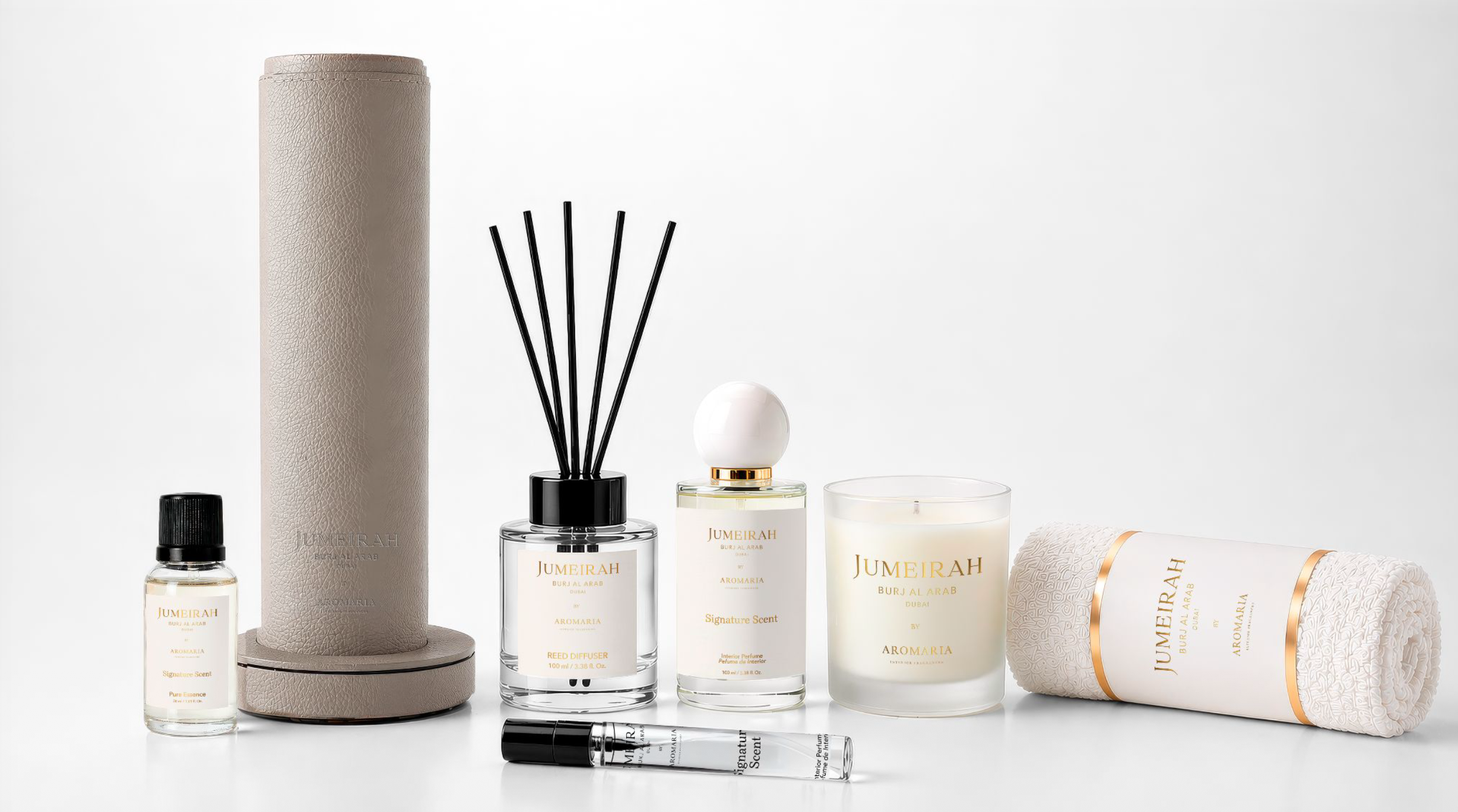 The full Jumeirah × Aromaria signature scent product lineup &mdash; essential oil vial, leather-wrapped diffuser cylinder, reed diffuser, signature scent bottle, scent vial, candle, and oshibori roll &mdash; arranged together on a soft neutral background.